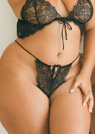 Woman wears a black lace thong with a bow in the front a thin side straps. The black thong is tied in the front with a bow and from Lioa Lingerie, a Swiss Lingerie Brand, made in Portugal.