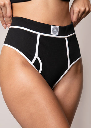 Woman wears a trendy sporty black high waist thong with a wide brand. The thong is inspired by boys underwear and features a lion head at the front center. This sporty lingerie is from Lioa Lingerie, a Swiss Lingerie Brand.